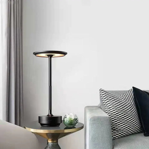 A stylish black table lamp with a circular LED light source, positioned on a gold-accented side table next to a grey sofa adorned with patterned cushions, creating a sophisticated and cozy living room ambiance.