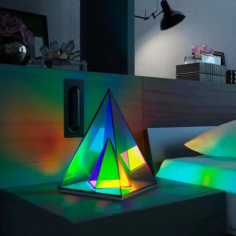 A colorful, triangular Infinity Lamp with a futuristic design, emitting vibrant LED lights, placed on a bedside table in a dimly lit room, creating a mesmerizing and modern ambiance next to a cozy bed.