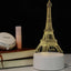 3D Hologram LED Lamp in the shape of the Eiffel Tower, featuring color-changing modes and a USB plug-in, placed on a desk beside books and a makeup compact.