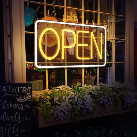 Open Vintage Neon Lamp with bright yellow LED lights displayed in a window, surrounded by flowers and creating a welcoming atmosphere for a business.